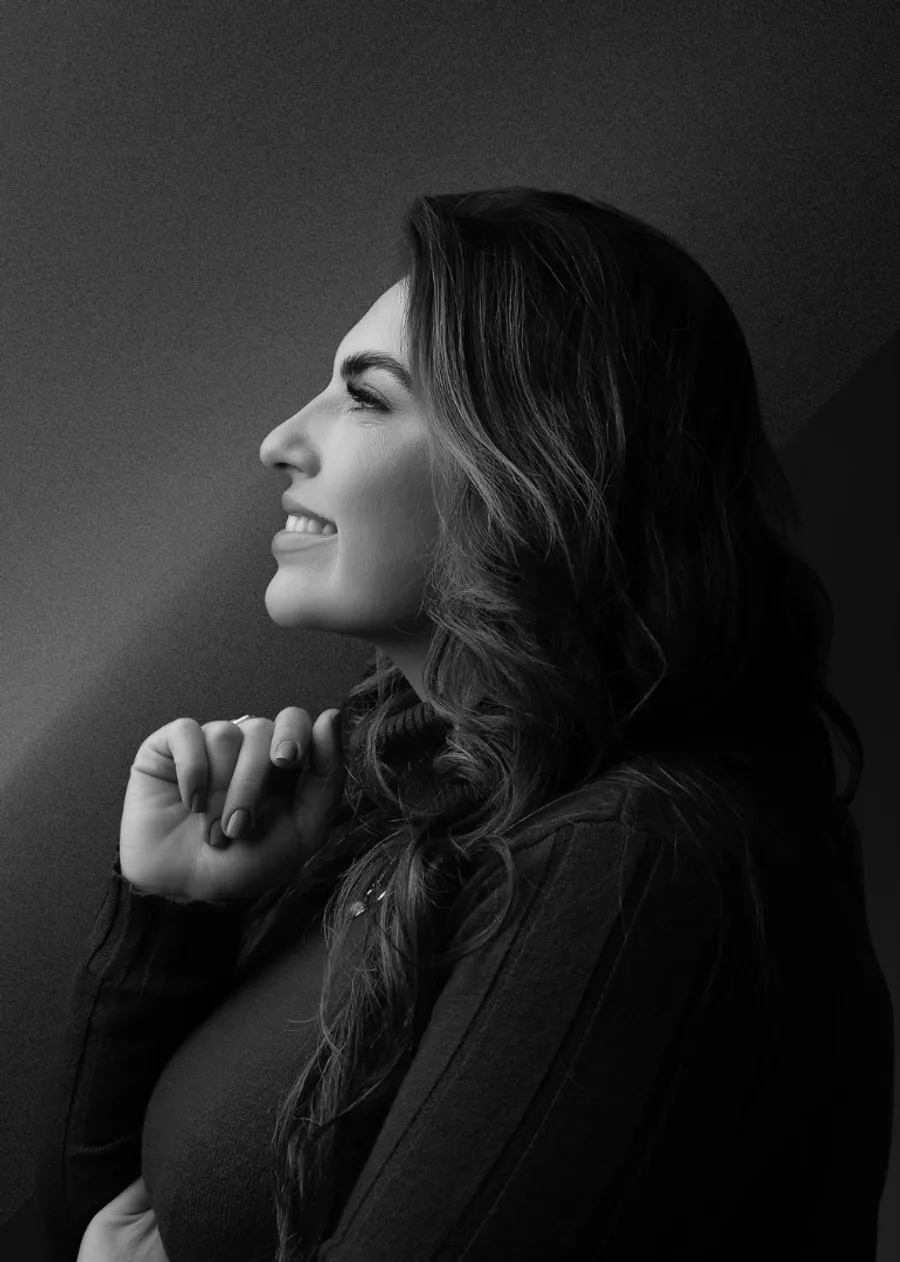 Tatiane Gracietti de perfil, em preto e branco, sorrindo com a mão no queixo